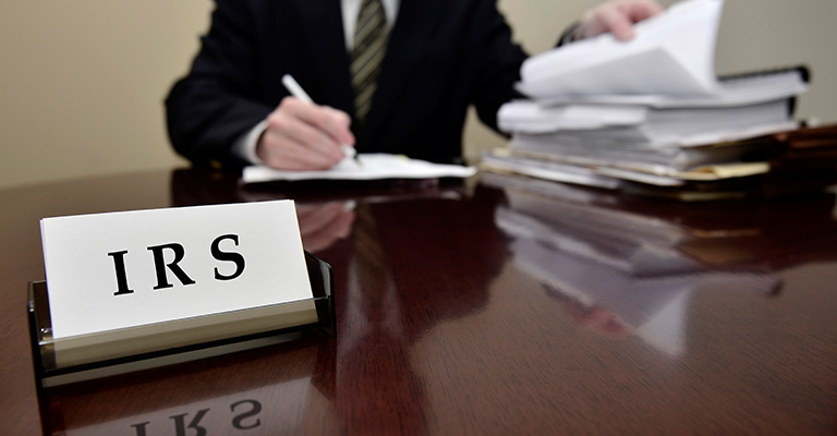 Man at desk with IRS nameplate, reviewing tax and appraisal documents, to symbolize proposed IRS regulations